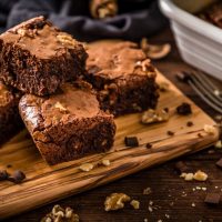 High angle view of various homemade chocolate brownie  slices on a wooden cutting board surrounded by some brownie crumbs, some walnuts, a fork and a baking sheet with a baked brownie on the inside. Predominant color is brown.  Studio shot taken with Canon EOS 6D Mark II and Canon EF 24-105 mm f/4L